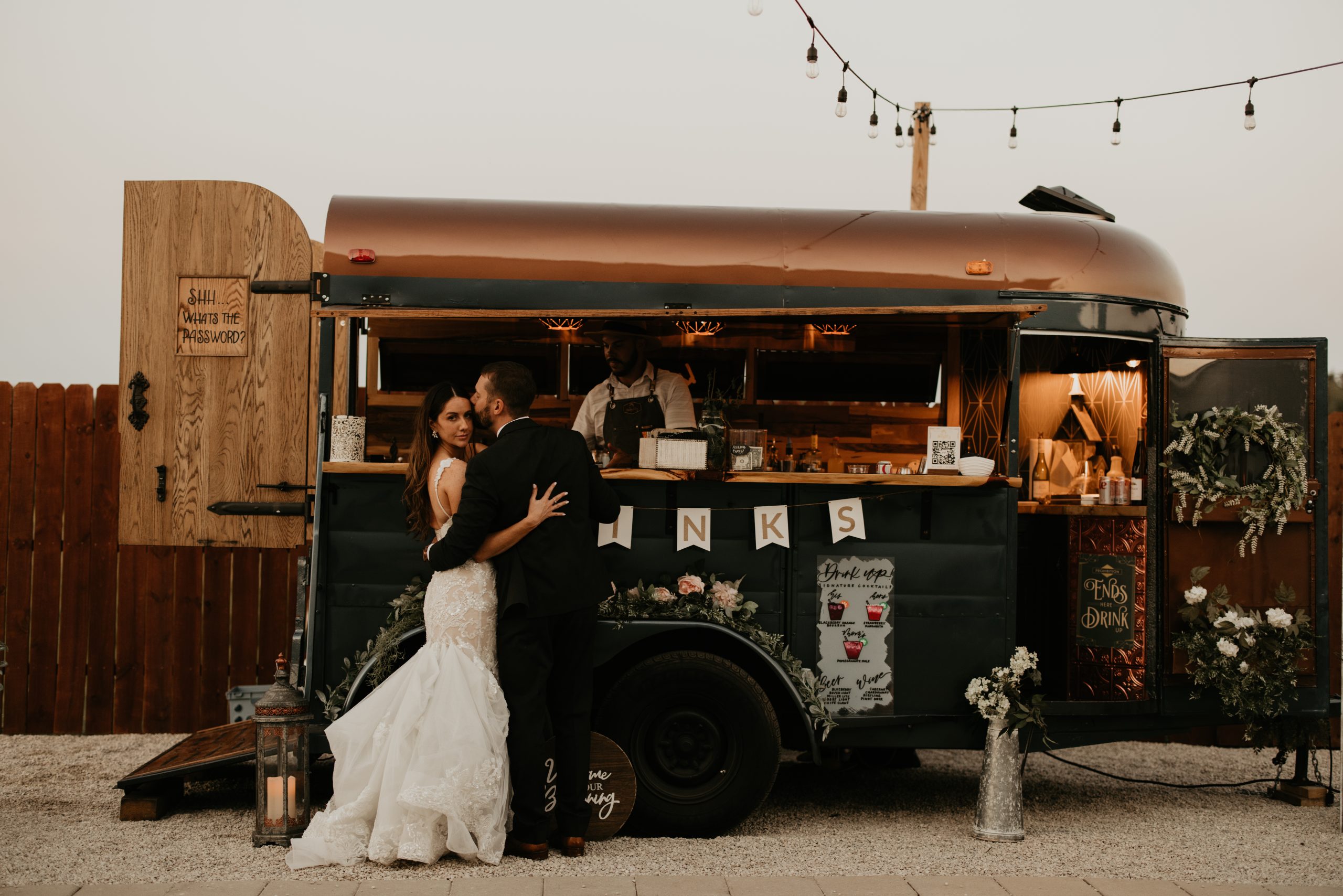 Bride and groom embracing in front of The Thirsty Filly's vintage horse trailer bar decorated with string lights and florals at sunset