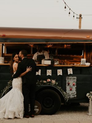Bride and groom embracing in front of The Thirsty Filly's vintage horse trailer bar decorated with string lights and florals at sunset