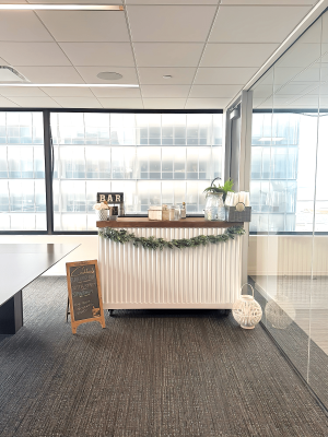 The Thirsty Filly's white corrugated-panel portable bar set up in an office breakroom with a 'BAR' sign and cocktail menu